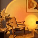 Lampe halo ambiance Golden Hour Dans un salon cosy, la lampe halo ambiance Golden Hour projette une lumière orange en cercle sur un mur, près d’un fauteuil en rotin et d’un bouquet de fleurs séchées.