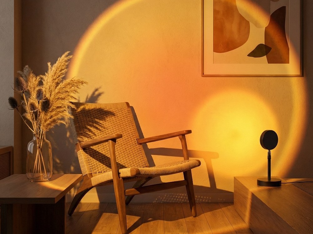 Dans un salon cosy, la lampe halo ambiance Golden Hour projette une lumière orange en cercle sur un mur, près d’un fauteuil en rotin et d’un bouquet de fleurs séchées.