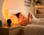 Une ado allongée dans un lit douillet utilise son téléphone, tandis que la lampe halo LED coucher de soleil USB projette un grand cercle lumineux orangé sur le mur.