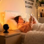 Une ado allongée dans un lit douillet utilise son téléphone, tandis que la lampe halo LED coucher de soleil USB projette un grand cercle lumineux orangé sur le mur.