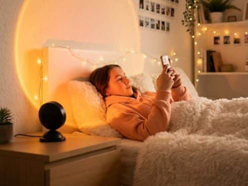 Une ado allongée dans un lit douillet utilise son téléphone, tandis que la lampe halo LED coucher de soleil USB projette un grand cercle lumineux orangé sur le mur.
