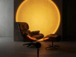 Un grand halo lumineux doré, projeté par la lampe halo LED sur pied effet coucher de soleil, éclaire un fauteuil design dans une pièce sombre et épurée.