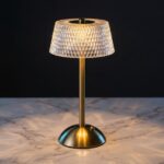 La lampe de table LED art déco rechargeable, abat-jour effet cristal et facettes diamant, pied brossé doré, posée sur un marbre clair.
