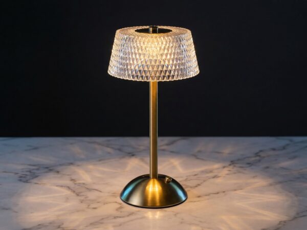 La lampe de table LED art déco rechargeable, abat-jour effet cristal et facettes diamant, pied brossé doré, posée sur un marbre clair.